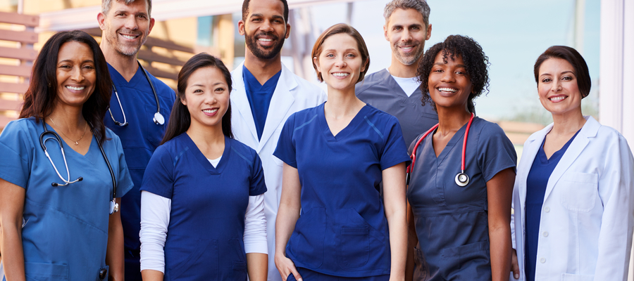 A group of nurses in blue scrubs and white jackets stood together smiling